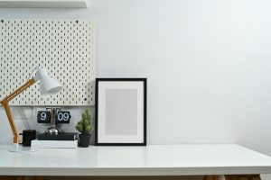 Image of blank picture frame, books, lamp, and vintage flip clock on white table against wall