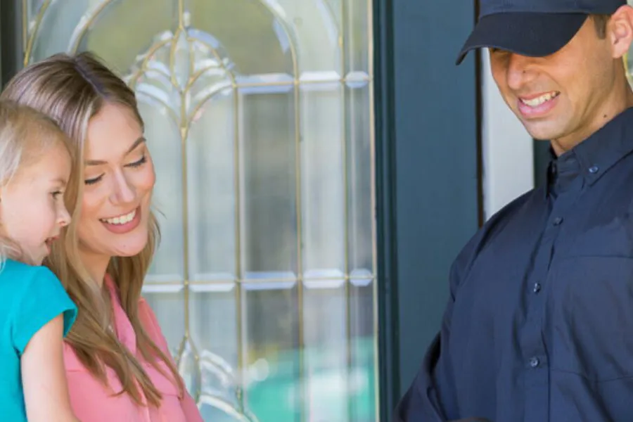 A friendly pest control professional in a dark uniform and cap smiling while speaking with a mother and her young daughter at the front door of their home.