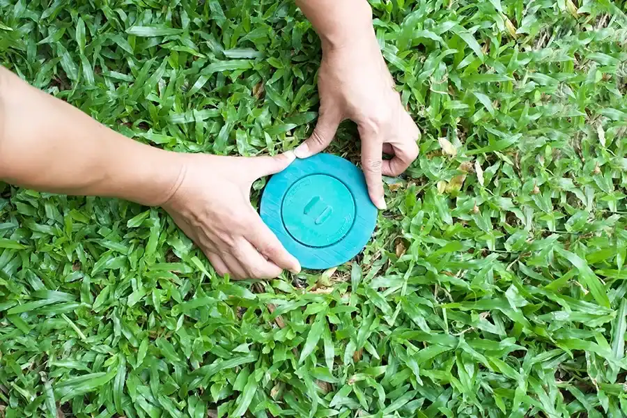 Two hands carefully placing a circular green plastic termite bait station lid into a hole in a lush green lawn.
