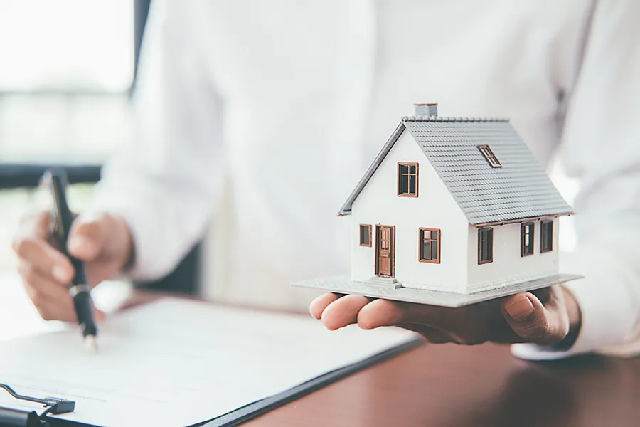 A person holding a small model of a white house in their palm while holding a pen over insurance paperwork on a clipboard, symbolizing home protection and policy coverage.