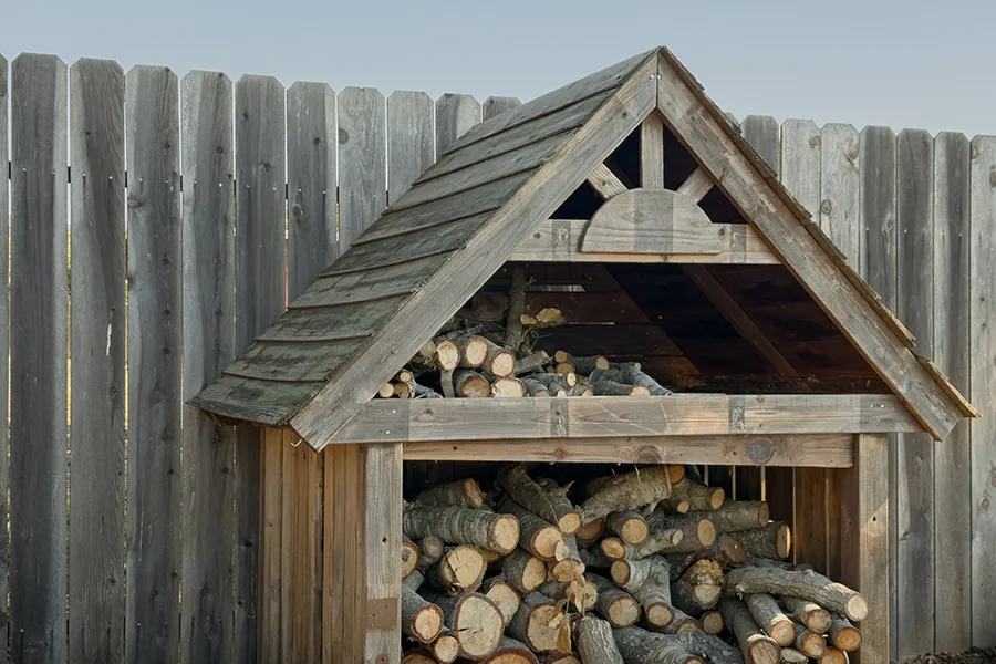 A wooden outdoor firewood storage shed filled with cut logs, standing against a grey wooden privacy fence.