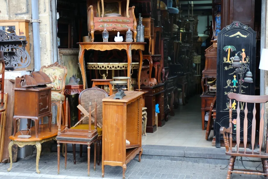 A variety of vintage and used wooden furniture, including chairs and tables, displayed on the sidewalk outside a secondhand store.