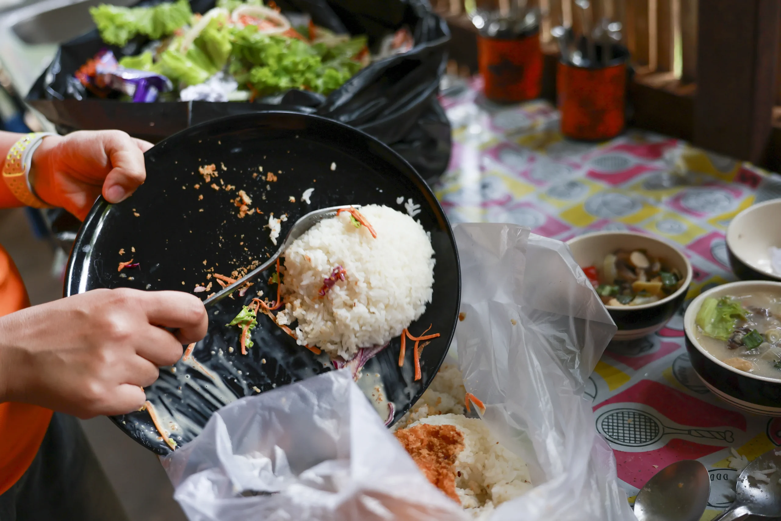 A person scraping leftover rice and food scraps from a black plate directly into a plastic garbage bag.
