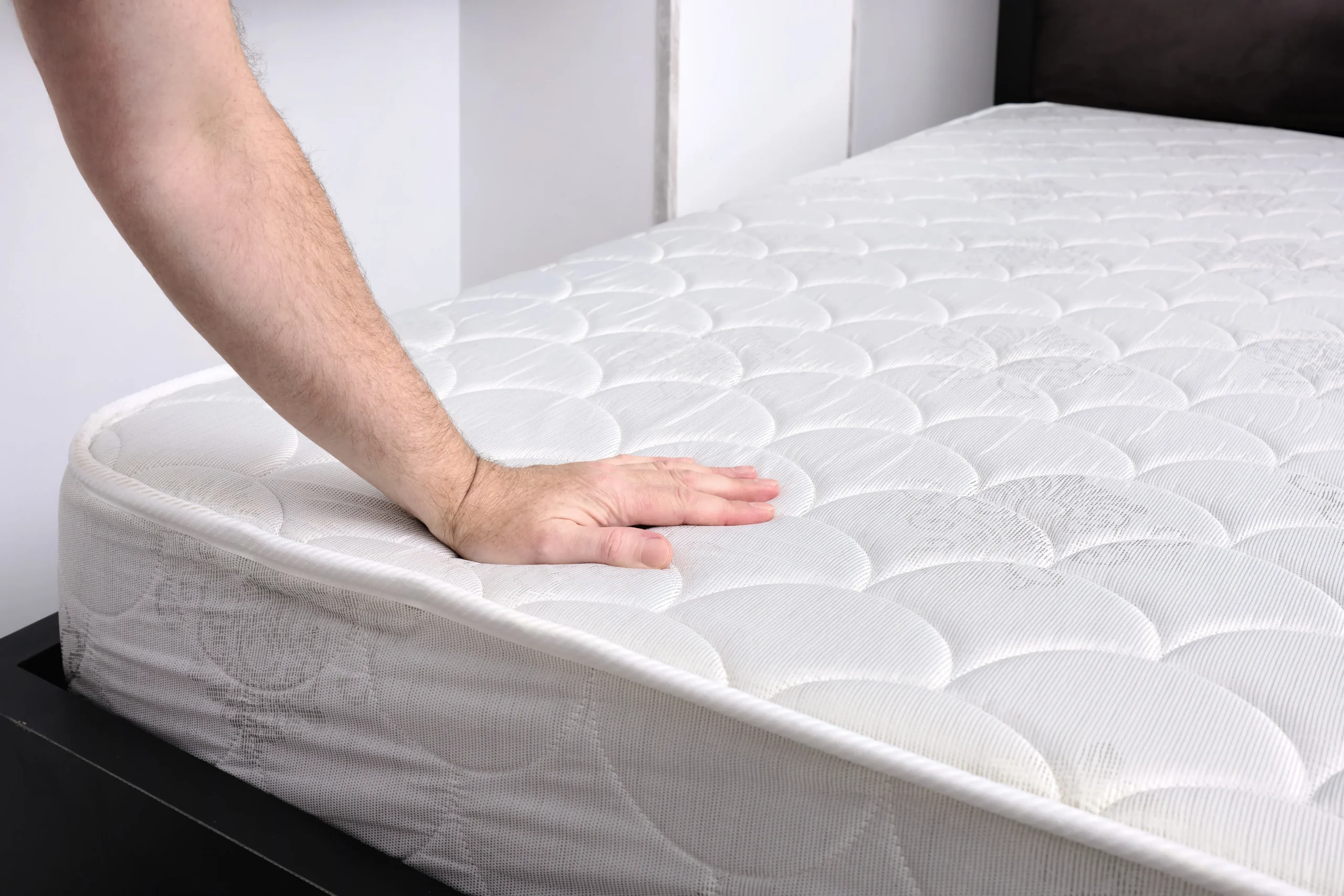 A person's hand pressing down firmly on a bare, white mattress resting on a dark bed frame.