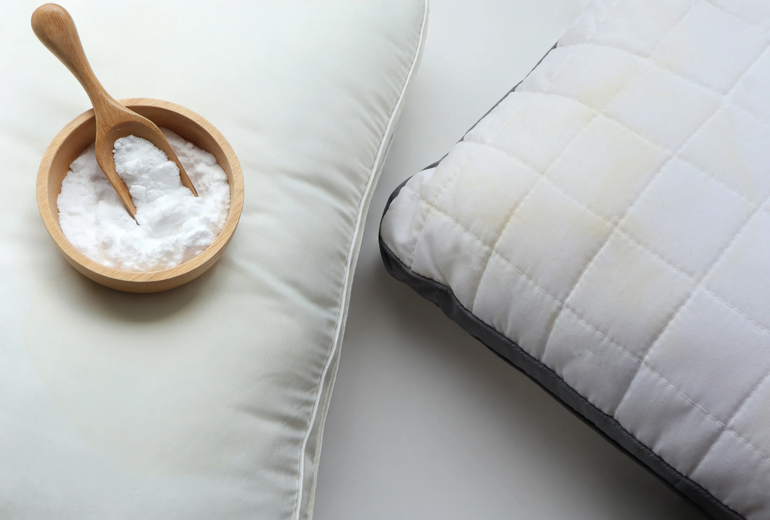 A wooden bowl of baking soda with a small wooden scoop resting next to a white quilted pillow.