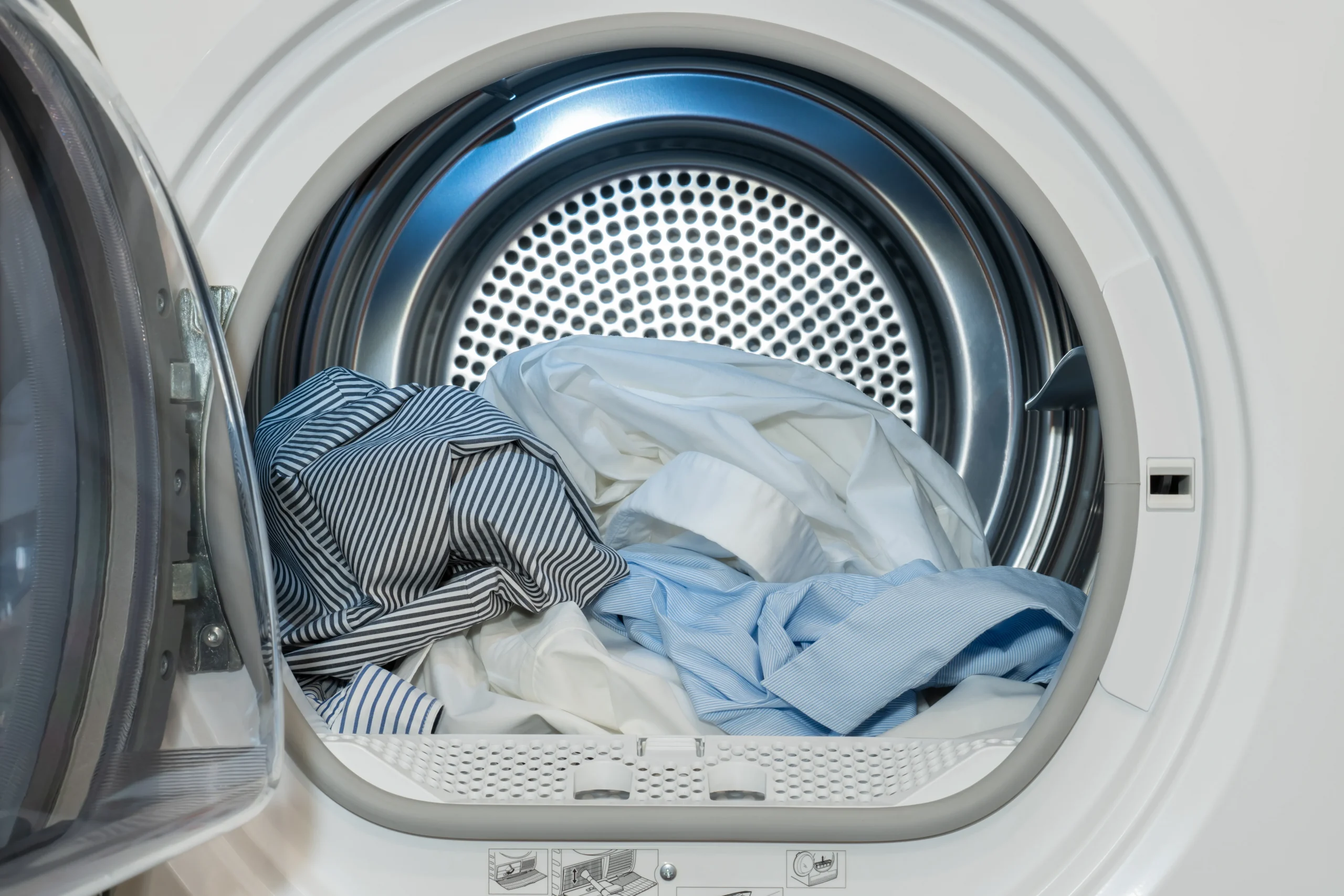 An open front-loading dryer filled with light-colored dress shirts and bedding.