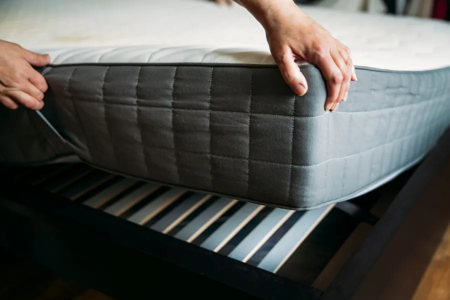Hands lifting a mattress protected by a grey zippered encasement off a slatted wooden bed frame.