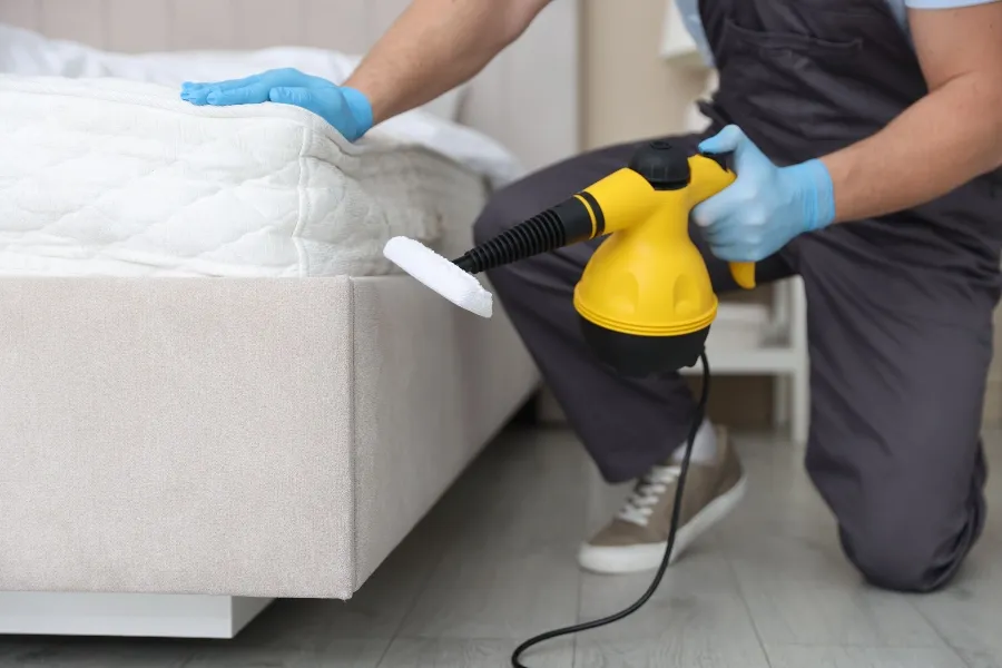 A pest control professional wearing blue gloves using a yellow handheld steam cleaner to treat the base of a mattress.