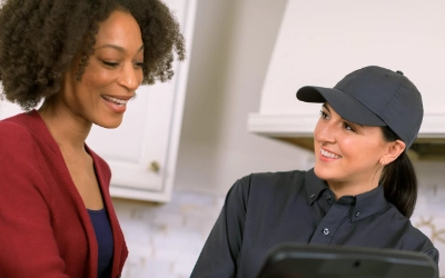 A smiling female pest control technician in a black uniform and cap speaks with a happy female homeowner in a kitchen.
